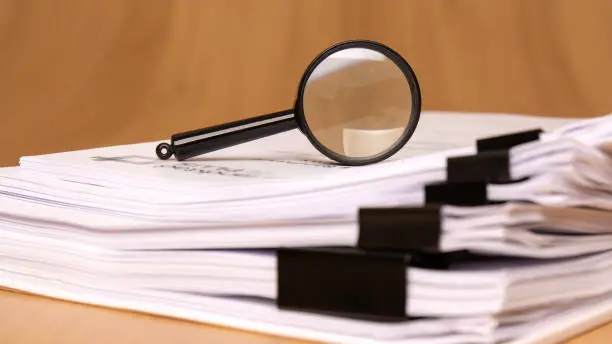 A magnifying glass resting on top of organized, stacked paper files held together by black binder clips, representing the required tenancy paperwork, Emirates ID copies, and municipal clearances needed for building management approval.