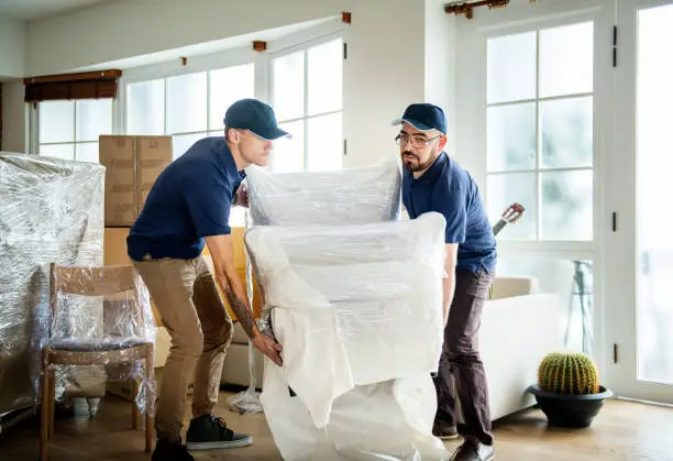 Two male movers carefully lifting a plastic-wrapped armchair, highlighting how premium protective packaging influences the total storage unit cost in Sharjah.