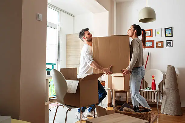 A young couple carrying a large, heavy cardboard box together inside their bright apartment, representing the official documentation and community clearance required by developers before tenants can legally transport household items in or out of a building.