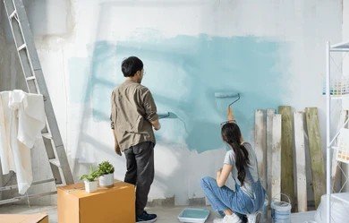 Two people painting an interior room wall light blue with paint rollers next to a ladder and drop cloths.